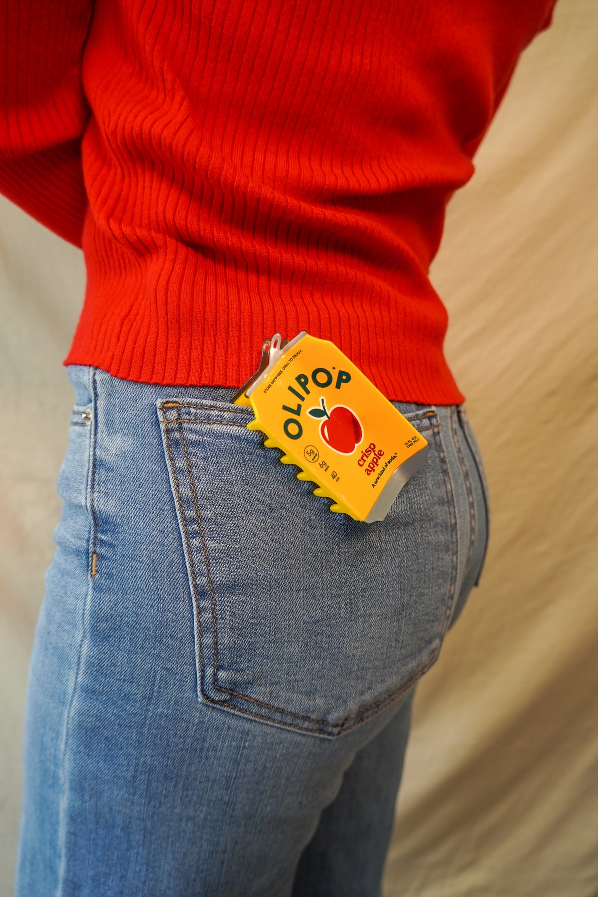 A close-up lifestyle shot of a person wearing a bright red ribbed sweater and light blue jeans, with a OLIPOP Crisp Apple Hair Claw clipped onto the back jean pocket.