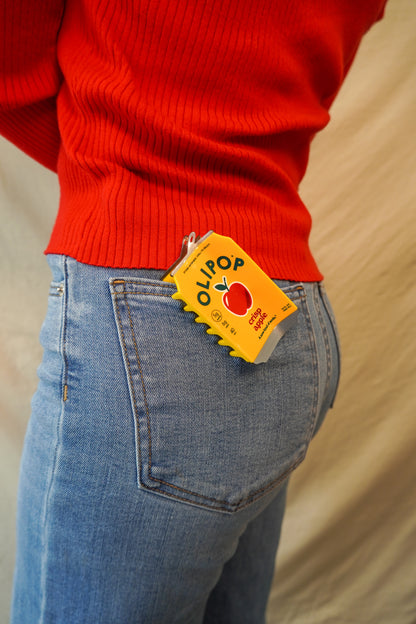 A close-up lifestyle shot of a person wearing a bright red ribbed sweater and light blue jeans, with a OLIPOP Crisp Apple Hair Claw clipped onto the back jean pocket.
