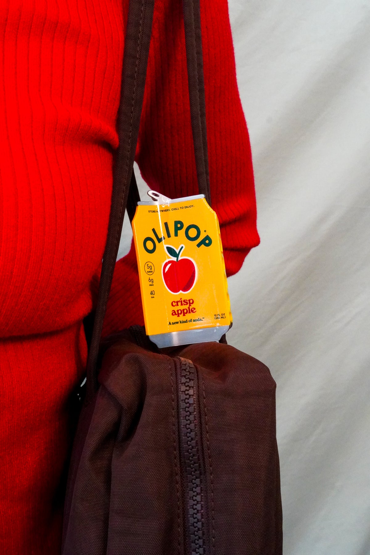 A close up of an OLIPOP crisp apple hair claw attached to the strap of a brown purse, being worn by a person in a red sweater