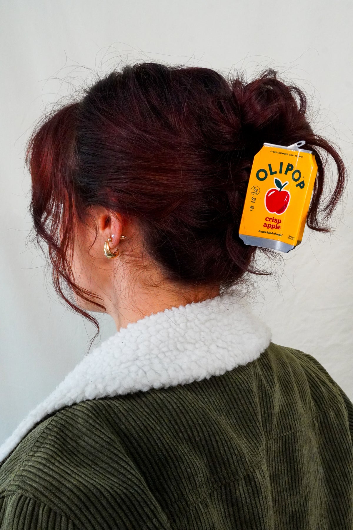 A person shown from the back wearing a dark olive green corduroy jacket with a white sherpa collar, with their hair in a bun secured by a OLIPOP Crisp Apple Hair Claw