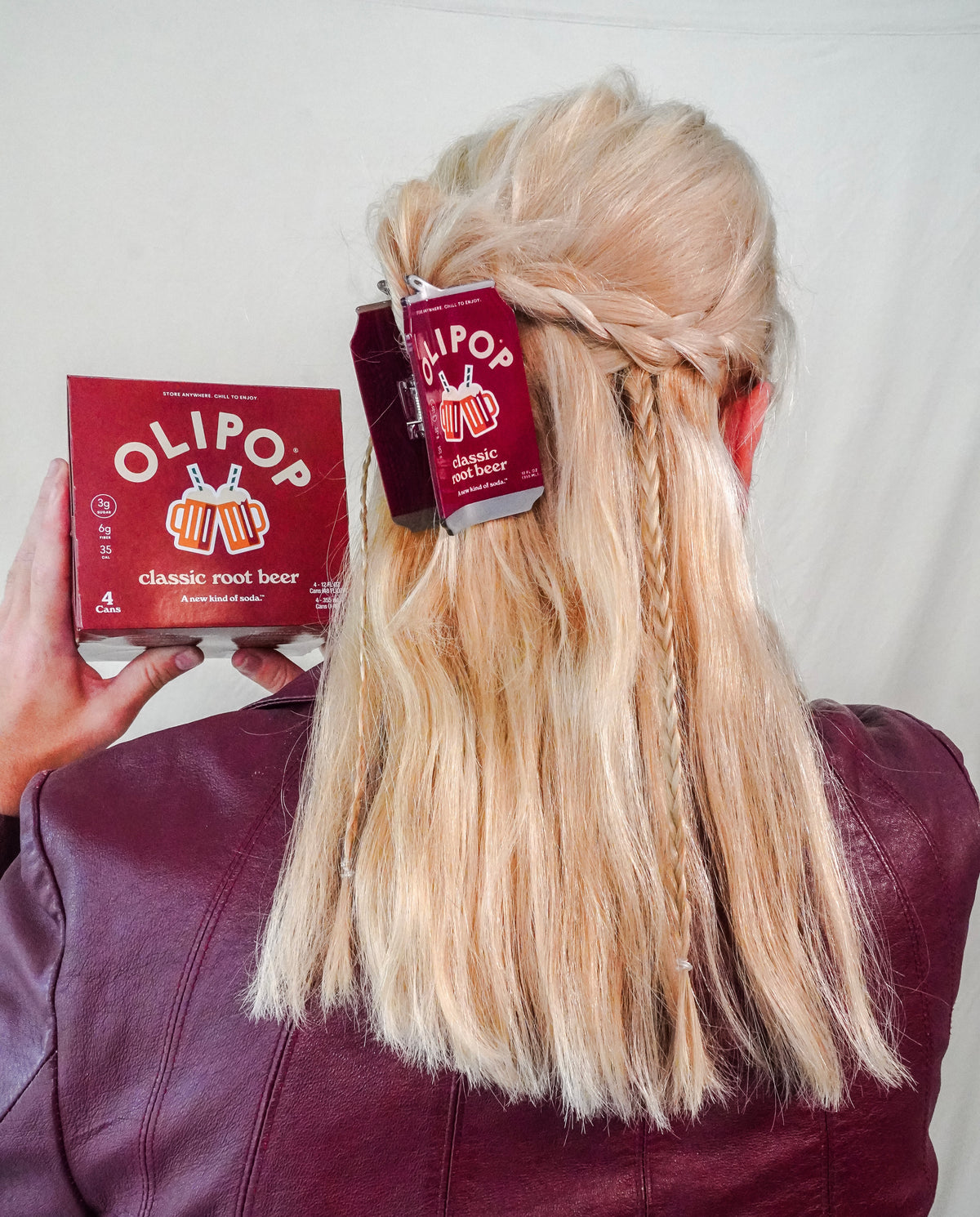 A person shown from behind wearing a maroon leather jacket, with long blonde hair styled in a half-up braid. They are holding a OLIPOP classic root beer box in their left hand, with a matching OLIPOP classic root beer hair claw holding the hair