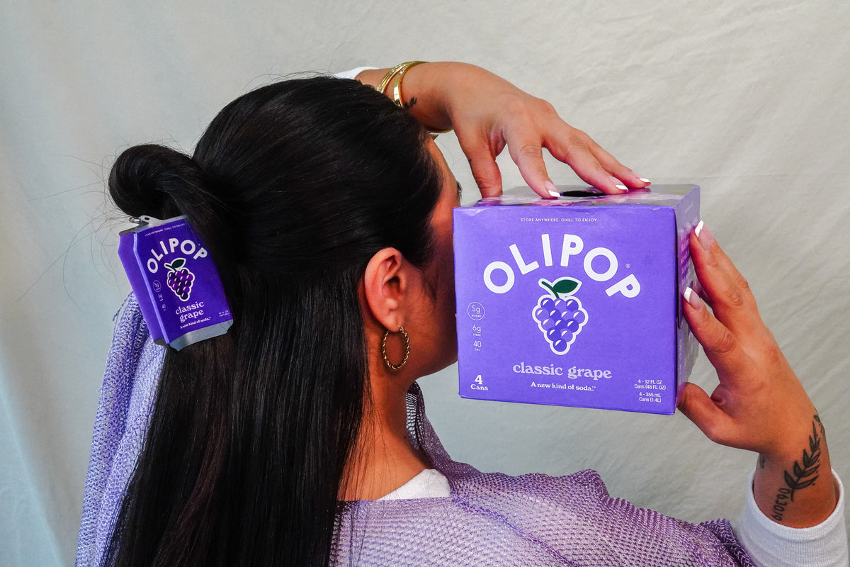 A woman holds a purple OLIPOP grape soda box next to her hair, which is styled with a matching Olipop Classic Grape hair claw. 