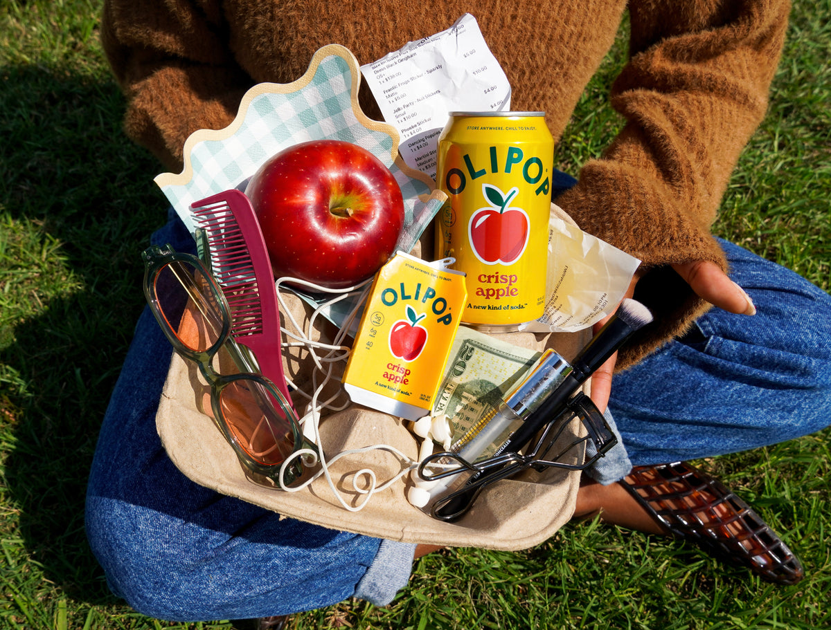 A person sitting outdoors on grass holds a tray filled with assorted items, including a red apple, sunglasses, earbuds, a pink comb, a Crisp Apple Olipop can, a Olipop Crisp Apple Hair claw, a receipt, and cash.