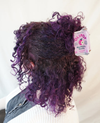 A person shown from the back wearing a white ribbed sweater and denim jacket, with curly hair in deep purple and black tones. A Olipop Blackberry Vanilla hair claw is holding their hair half up