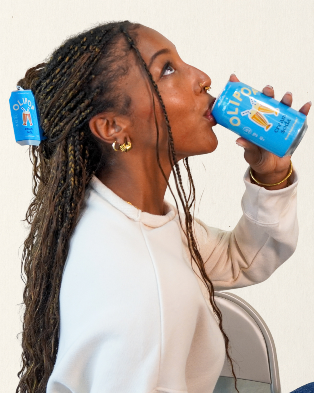 A woman with long braided hair drinks from a blue can of Olipop Cream soda, with a Olipop Cream Soda hair claw in her hair