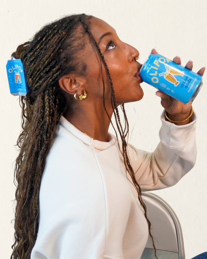 A woman with long braided hair drinks from a blue can of Olipop Cream soda, with a Olipop Cream Soda hair claw in her hair
